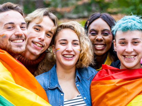 A group of people smiling and have a rainbow flag wrapped around them.