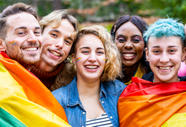 A group of people smiling and have a rainbow flag wrapped around them.