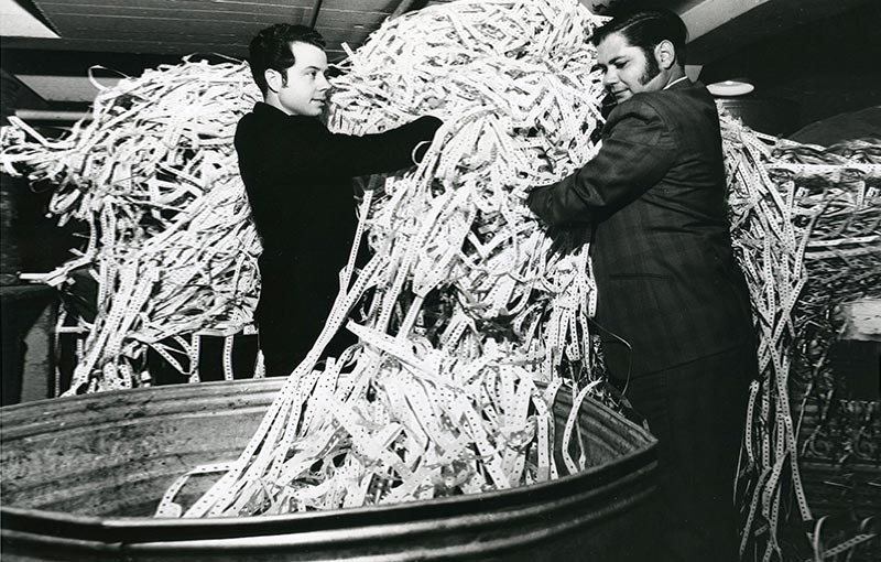 Two men in suits handling a large tangled pile of punched tape as part of manual data management.