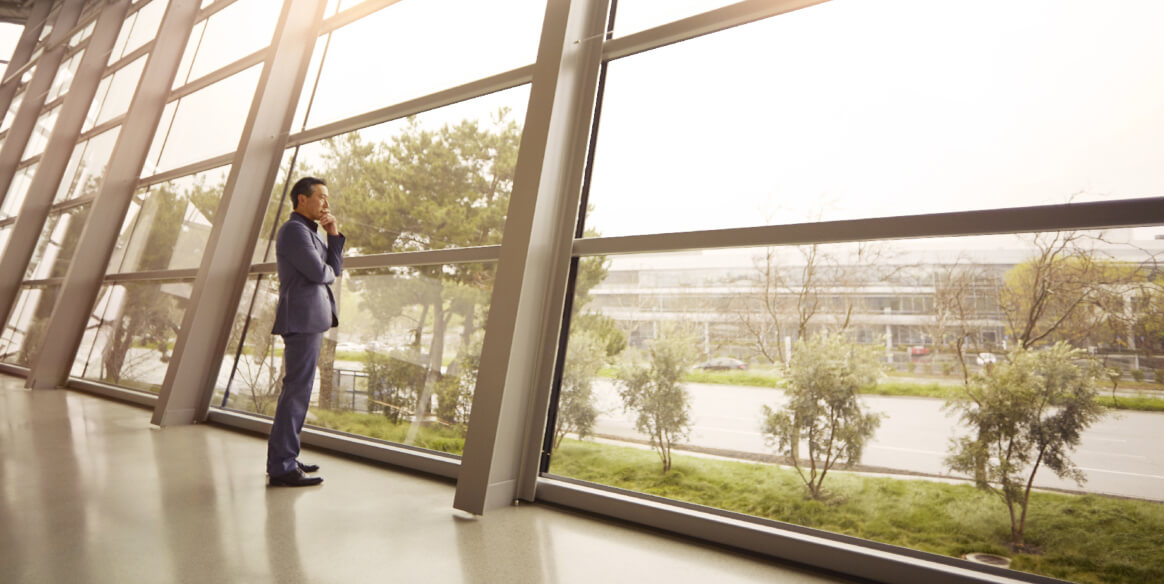 A person gazes through a wall of glass windows at an office courtyard.