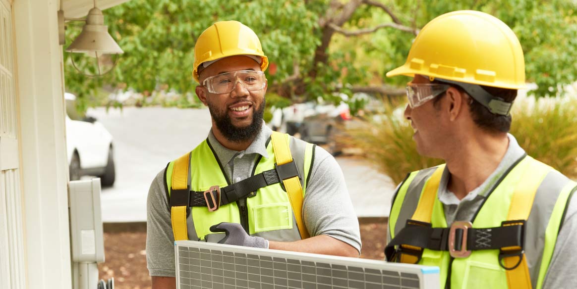 Two workers in hard hats and safety goggles install a solar panel.